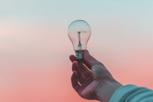 A hand holding up a lightbulb against a gradient backdrop.